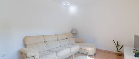 A modern living room with a sectional sofa, a glass coffee table, a small side table, a lamp, a potted plant, and a TV stand.