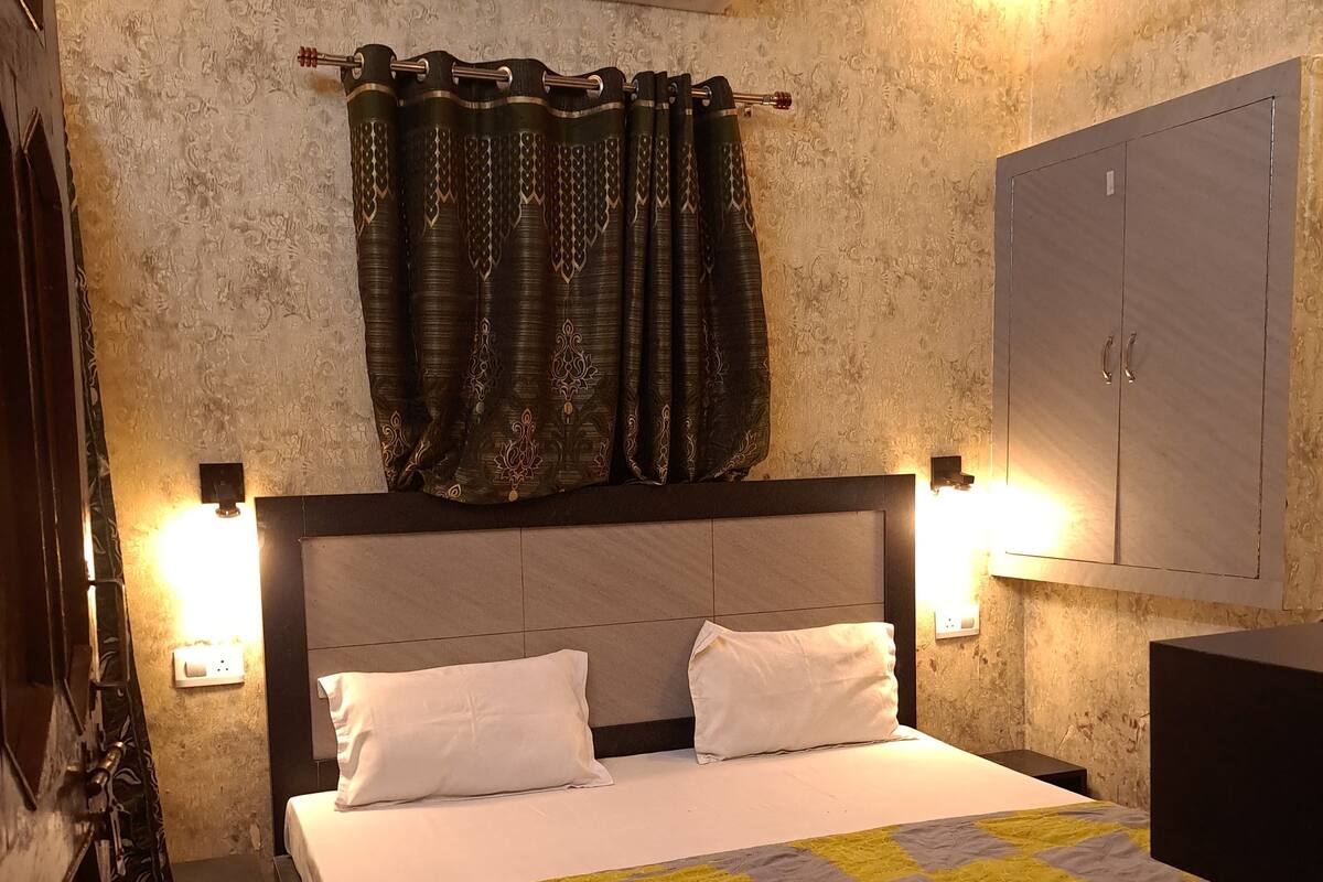 A bedroom with a bed, two bedside lamps, a wall-mounted air conditioner, and patterned curtains.