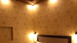 A neatly made bed with a patterned bedspread and two pillows, a headboard with a light fixture, and a textured wall with a recessed ceiling light. - Shree Ji Inn Homestay (Rishikesh)