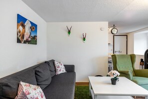 Una sala de estar con un sofá gris, un sillón verde, una mesa de centro blanca y un cuadro de una vaca en la pared.
