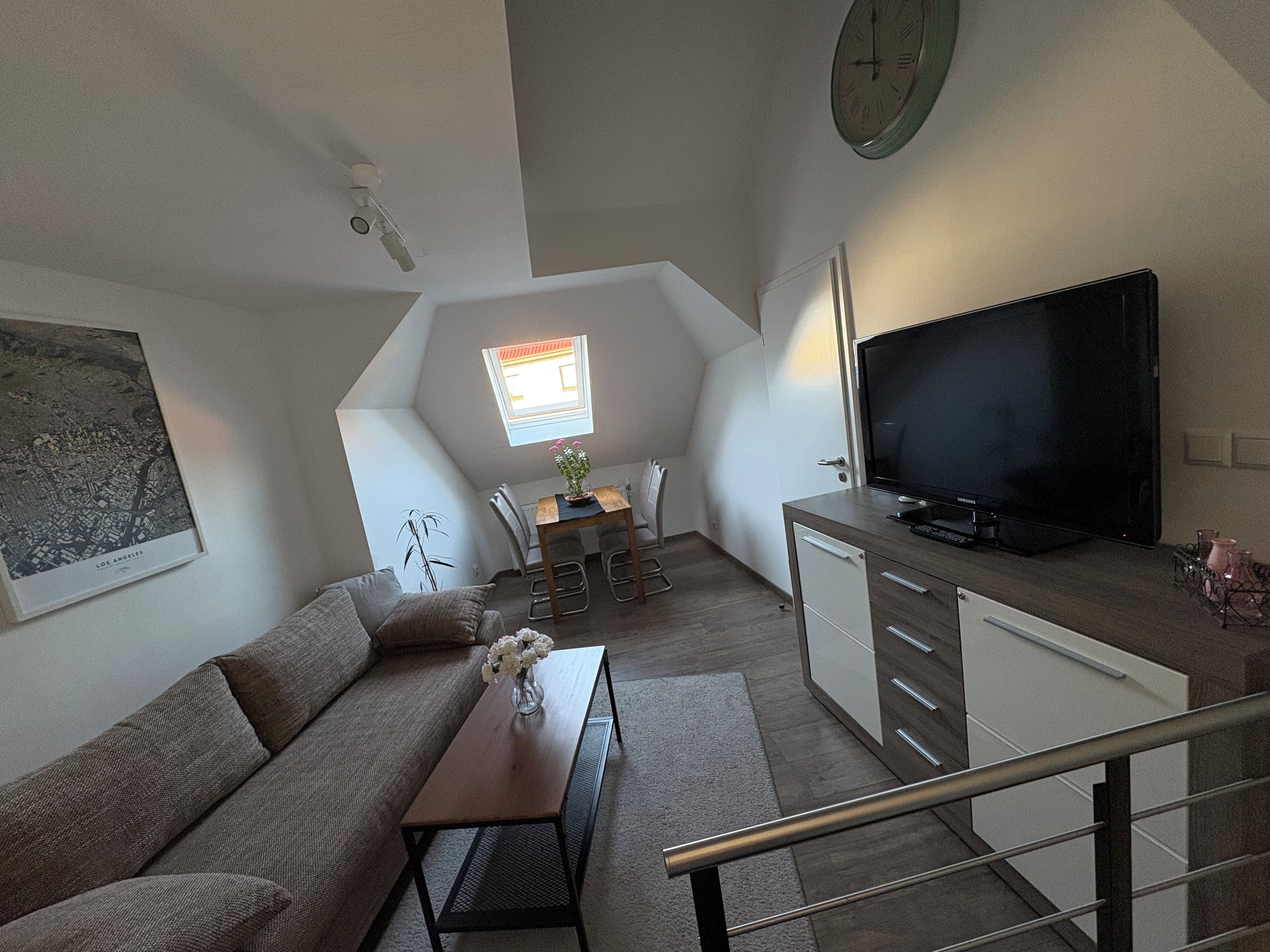 Un espacio habitable compacto con sofá, una pequeña mesa de comedor con sillas, televisor y un reloj en la pared.