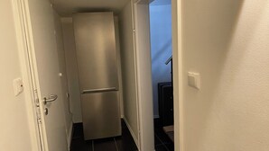Ein schmaler Flur mit Kühlschrank und Tür links, Lichtschalter rechts und einem dunklen Fliesenboden.