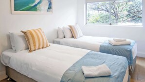Una habitación con dos camas, cada una con una almohada y una manta. Hay una ventana que da a árboles y un cuadro en la pared.