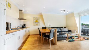 A modern kitchen with white cabinets, a dining area with a wooden table and chairs, a gray sofa, and a large window with a view. - Apartment 'Duhnen' with Balcony and Wi-Fi (Cuxhaven)