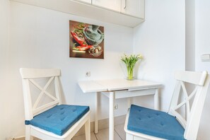 Un petit coin repas avec deux chaises blanches dotées de coussins bleus, une table blanche et un tableau accroché au mur représentant un mortier et un pilon, avec des poivrons rouges.