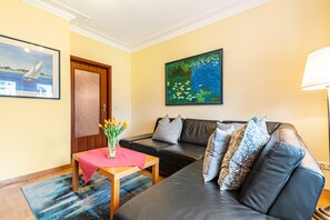 Un salon avec un canapé, une petite table sur laquelle est posé un vase de fleurs, un tableau au mur et une porte.