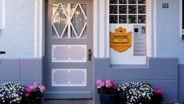 A building facade with a door, a window with a geometric pattern, a mailbox, and flower pots.