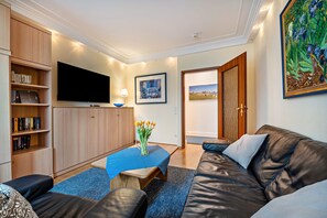Ein modernes Wohnzimmer mit einem Flachbildfernseher, Holzregalen, einem blauen Tisch mit Blumen in einer Vase und einem Ledersofa.