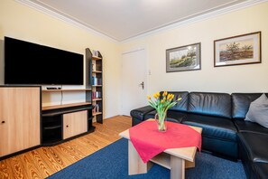 Una sala de estar con una televisión de pantalla plana, una estantería, un sofá de cuero negro, una mesa roja con un jarrón de flores y dos cuadros en la pared.