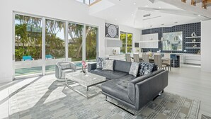 A modern living room with a grey sofa, a coffee table, and a large window overlooking a pool. - Luxury New Modern Beach Mansion in BEST Location! (Jupiter)
