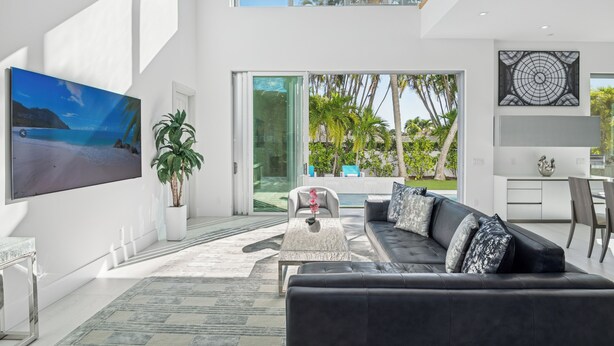 A modern living room with a large flat-screen TV, a black leather sofa, a coffee table, and a potted plant. - Luxury New Modern Beach Mansion in BEST Location! (Jupiter)