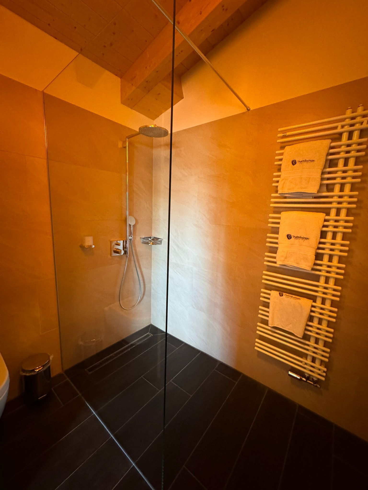 A modern bathroom with a glass shower enclosure, black tiled floor, and a towel rack with towels.
