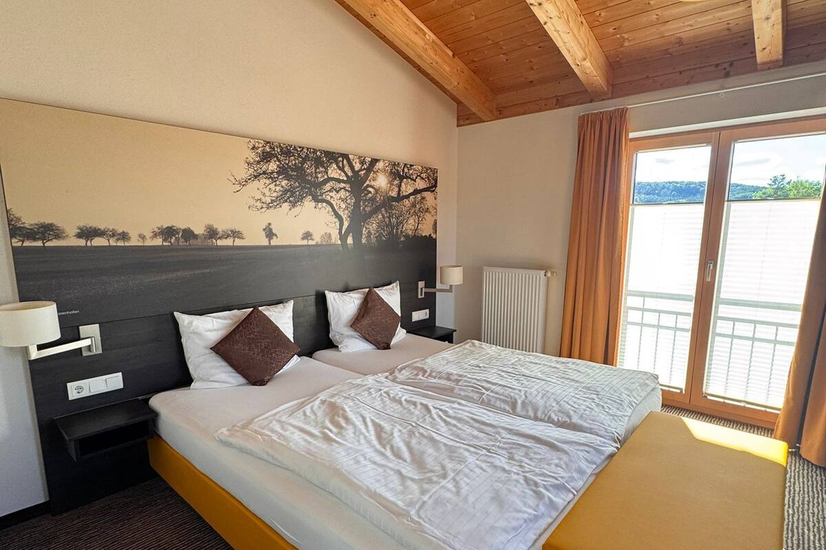 Ein Hotelzimmer mit Holzbalkendecke, einem großen Bett mit weißer Bettwäsche, einem Nachttisch, einer Lampe und Blick ins Freie durch ein Fenster.