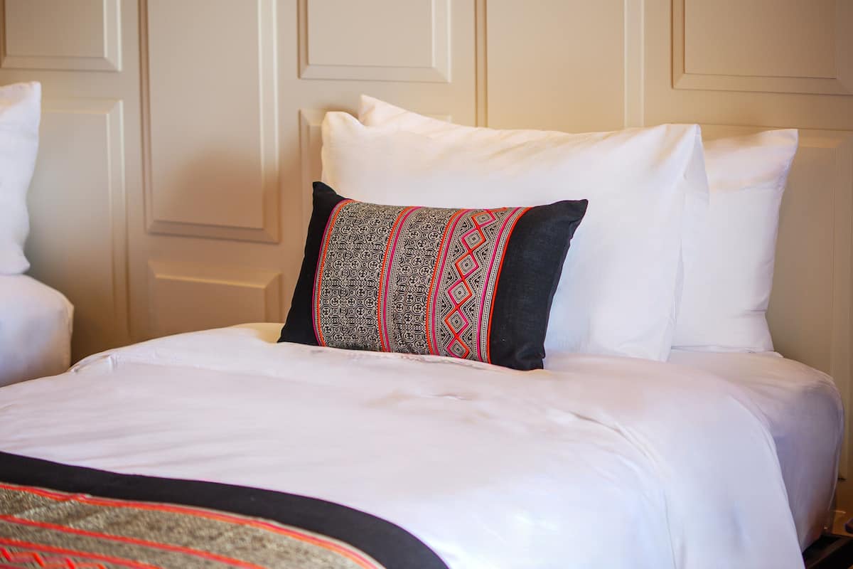 Un lit bien fait, avec du linge de lit blanc et un seul coussin décoratif orné d’un motif géométrique.