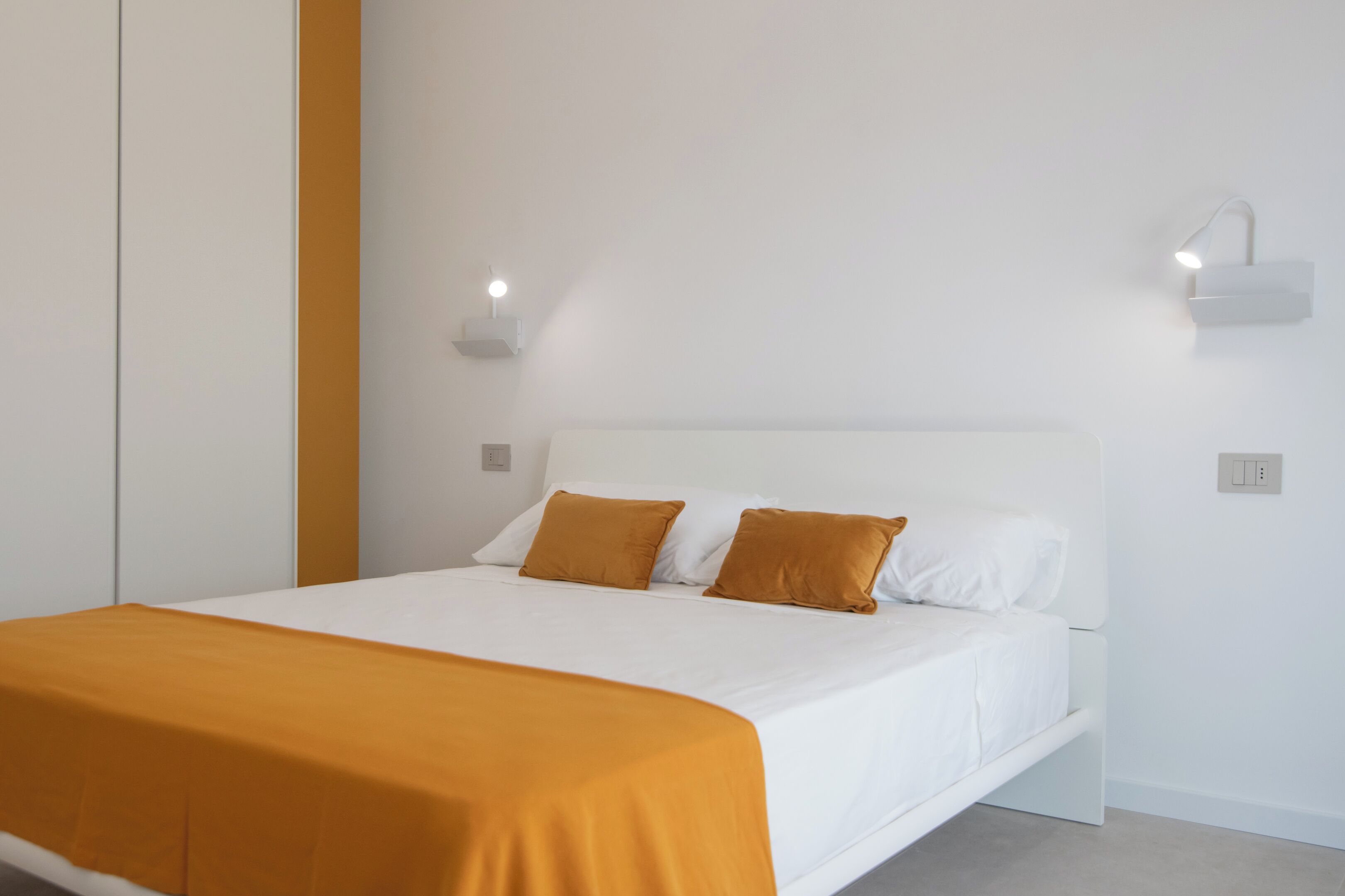 Une chambre à coucher moderne avec un lit blanc, deux oreillers marron et une tête de lit blanche. Deux lampes murales sont fixées de chaque côté du lit.
