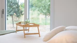 Un letto con biancheria e cuscini bianchi, un vassoio di legno con un bicchiere e una bottiglia, e vista sugli alberi attraverso ampie finestre.
