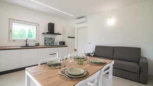 Una cucina moderna con mobili bianchi, un tavolo da pranzo in legno apparecchiato con piatti e bicchieri, e un divano grigio nella zona soggiorno.