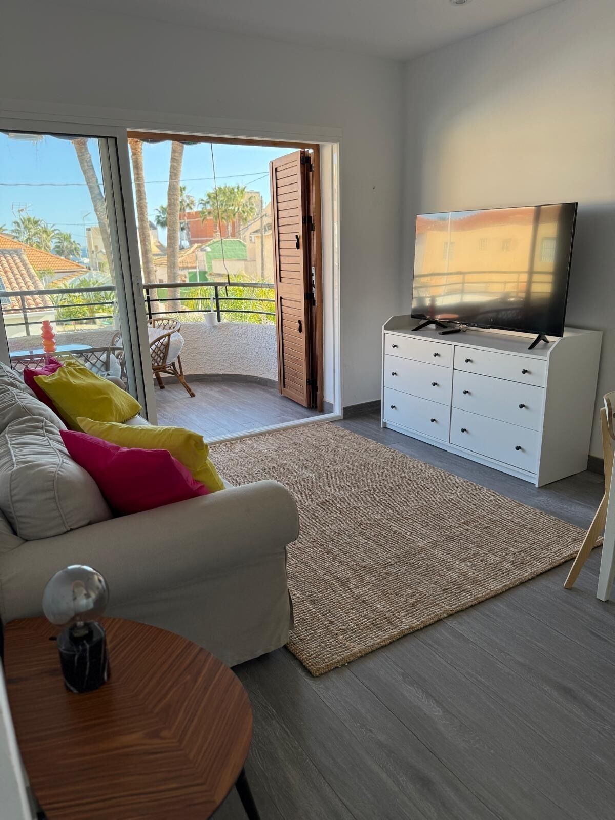Ein modernes Wohnzimmer mit einer Couch, einem Couchtisch, einem Fernseher auf einem weißen Schrank und einem Balkon mit Blick auf Palmen und Gebäude.