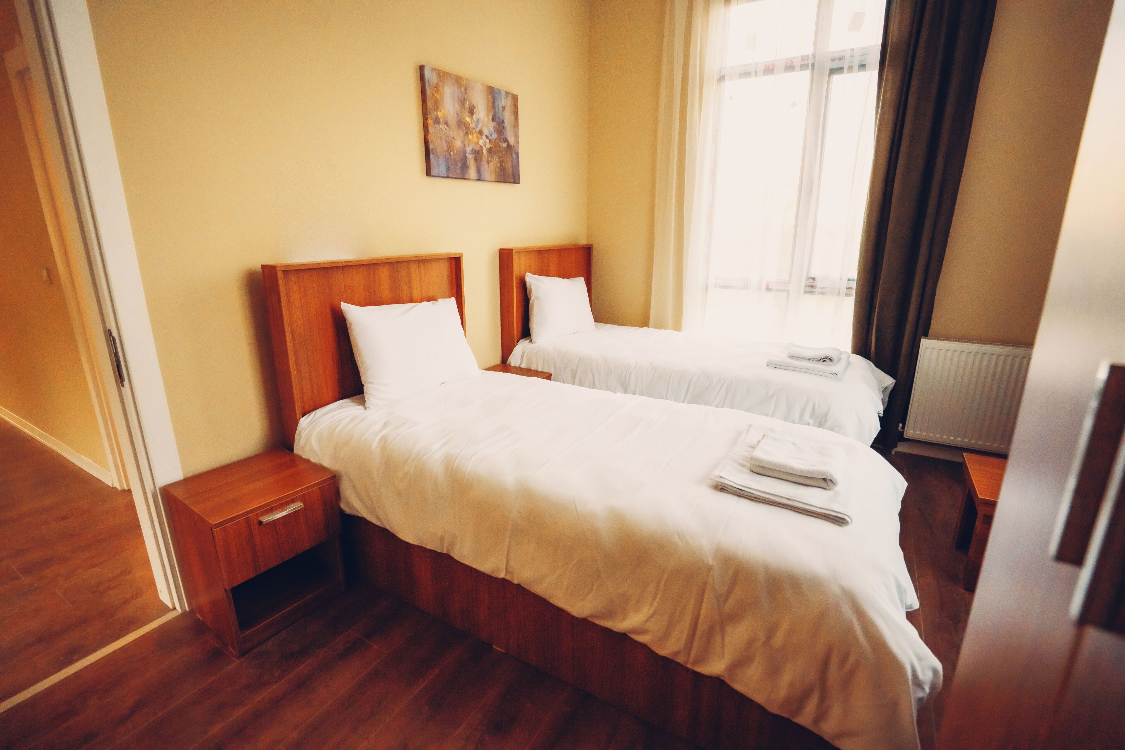 A hotel room with two beds, wooden headboards, white bedding, a small bedside table, a radiator, and a window with curtains.