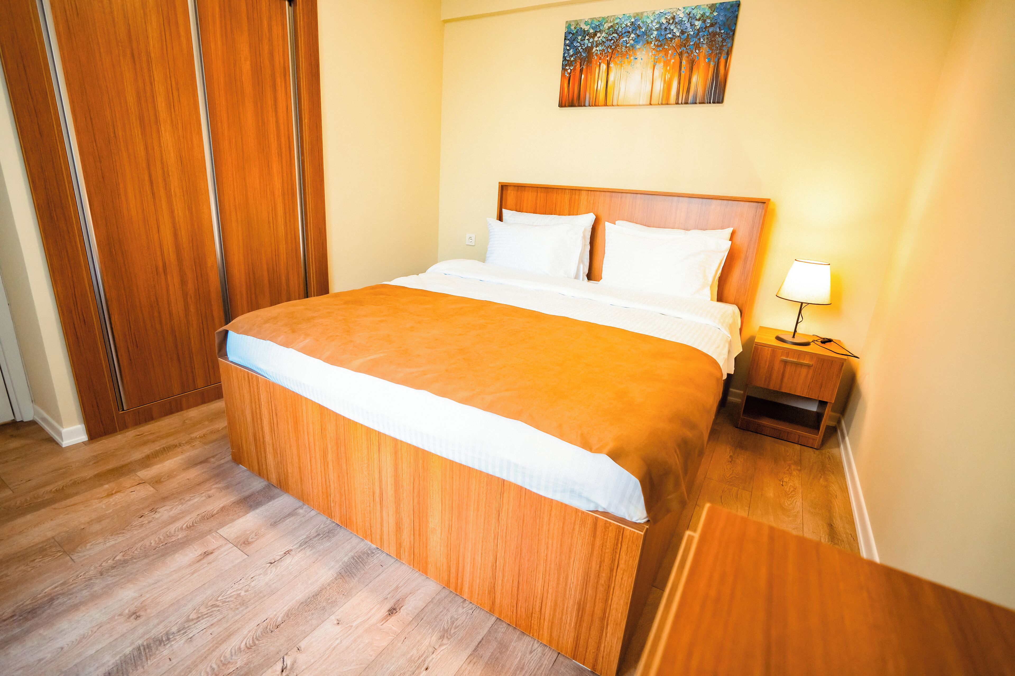 Een opgemaakt bed met houten hoofdbord, een nachtkastje met lamp en een schilderij aan de muur.
