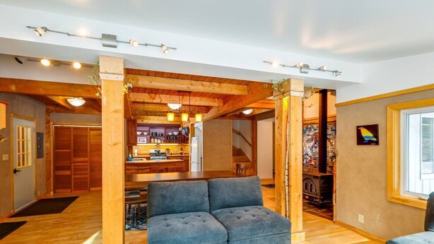A cozy living room with a sofa, wooden beams, a kitchen in the background, and a staircase.