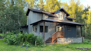 A two-story house with a wooden staircase leading to a porch, surrounded by lush greenery and trees.