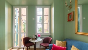 Een kamer met een blauwe bank, twee rode stoelen, een kleine ronde tafel met een vaas en een wijnglas, en een grote spiegel aan de muur.