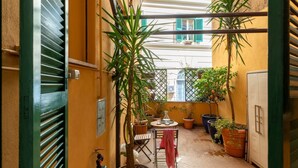 Een smalle, buitenste gang met potplanten, een tafel en stoelen, die leidt naar een balkon met uitzicht op gebouwen.