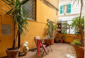 Pieni patio, jossa on pöytä ja tuoleja, ja jota ympäröivät ruukuissa olevat kasvit ja suuri puu.