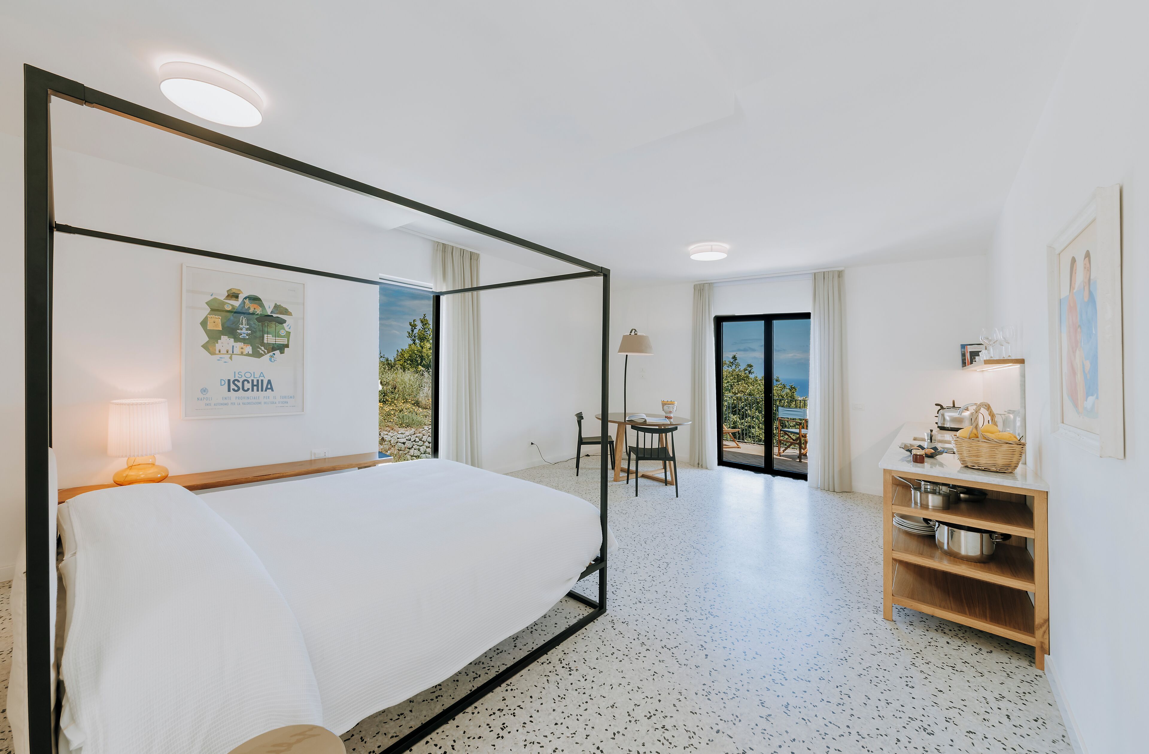 A modern hotel room with a large bed, a small table with a lamp, a wooden side table with a basket, and a view of the outdoors.