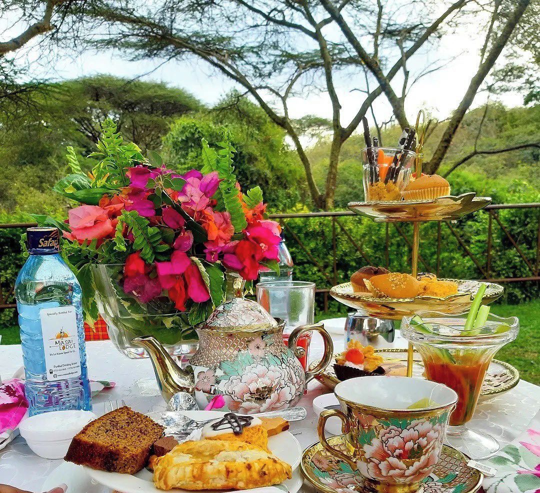 A table set with a bottle of water, a floral vase, a teapot, a tiered tray with pastries, and a cup with a saucer.