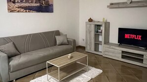 A modern living room with a grey sofa, a coffee table, a TV with a Netflix sign, a painting on the wall, and a cabinet.