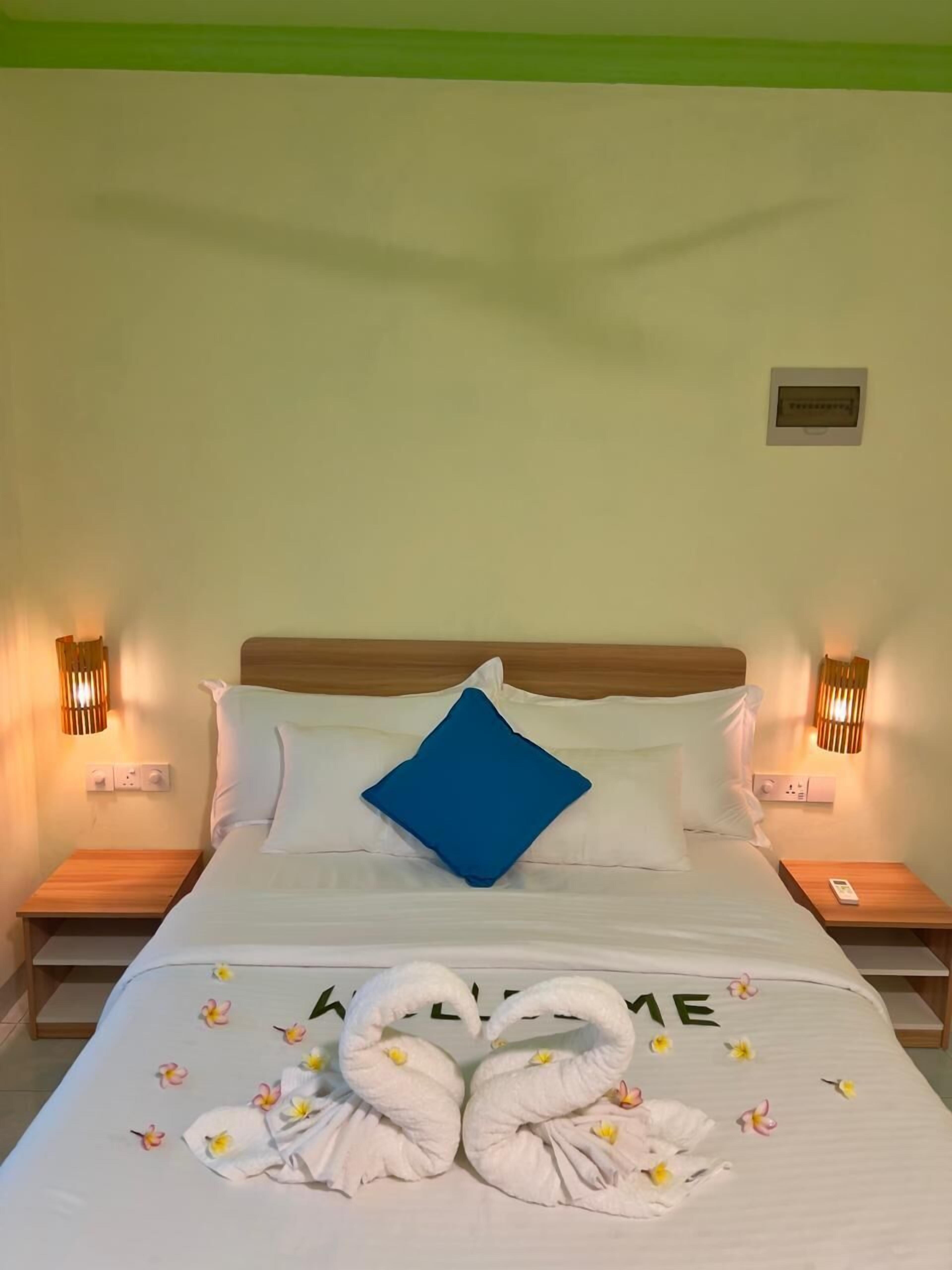 Una cama bien hecha con un almohábil azul, dos mesitas de noche con lámparas y un interruptor de luz en la pared.