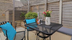 An outdoor patio with a metal table and chairs, a bottle of wine, and a vase with flowers. - Cedar Drive (Weymouth)