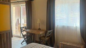 A bedroom with a bed, a small table with a lamp, a chair, and a bench. There is a window with curtains and a view of the outside.