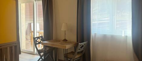A bedroom with a bed, a small table with a lamp, a chair, and a bench. There is a window with curtains and a view of the outside.
