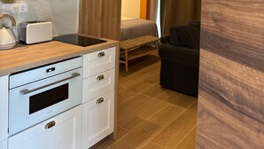 A modern kitchen with a built-in oven, a toaster, and a kettle. There is a wooden cabinet and a black range hood. A small table with a basket and a cup is visible. The room has a wooden floor and a gray sofa in the background. - ARENAS SUITES APARTAMENTOS (Potes)