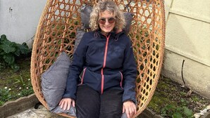 Une personne assise dans un fauteuil suspendu en osier, dans un cadre extérieur.