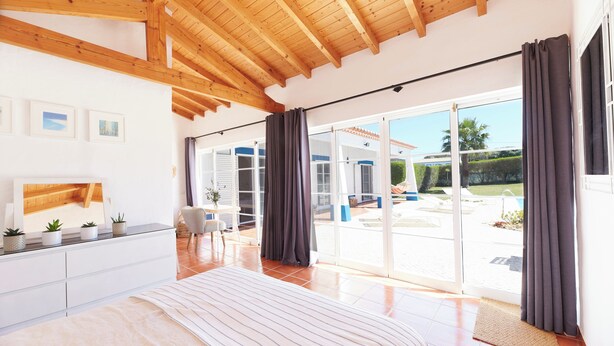 A spacious bedroom with a large bed, wooden ceiling beams, a sliding glass door to a patio, and a white dresser with potted plants. - Ocean Whisper-spectacular villa on Portugal's west coast (Aljezur)