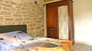 A bedroom with a stone wall, a wooden wardrobe, a bed with a floral bedspread, and a ceiling light fixture. - Spacious cozy and pet friendly Gîte (6pers with 3 bdrms/2 baths/3 toilets). (Claudon)