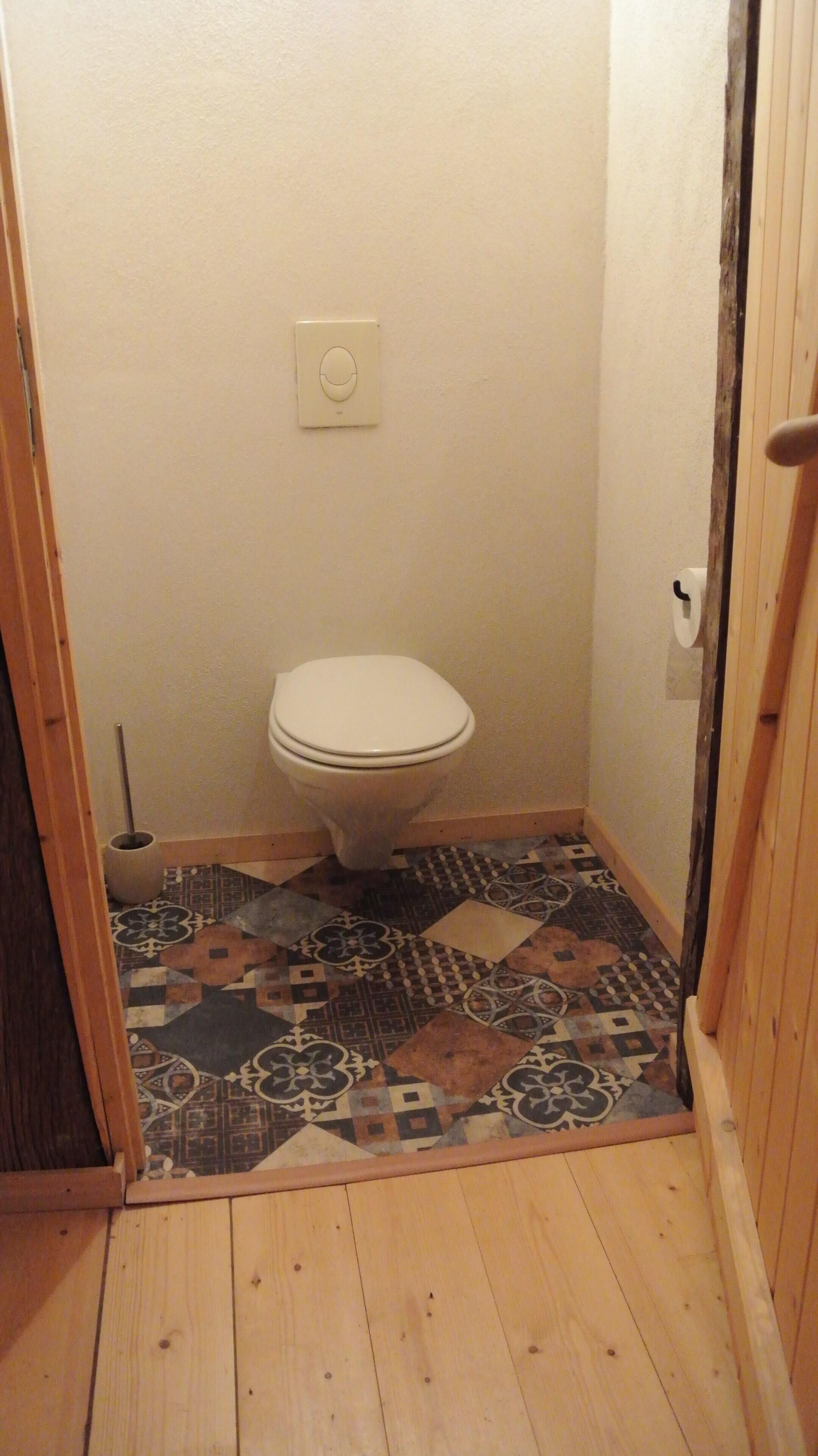 Un baño pequeño y estrecho con inodoro, dispensador de papel higiénico y suelo de baldosas con diseño.