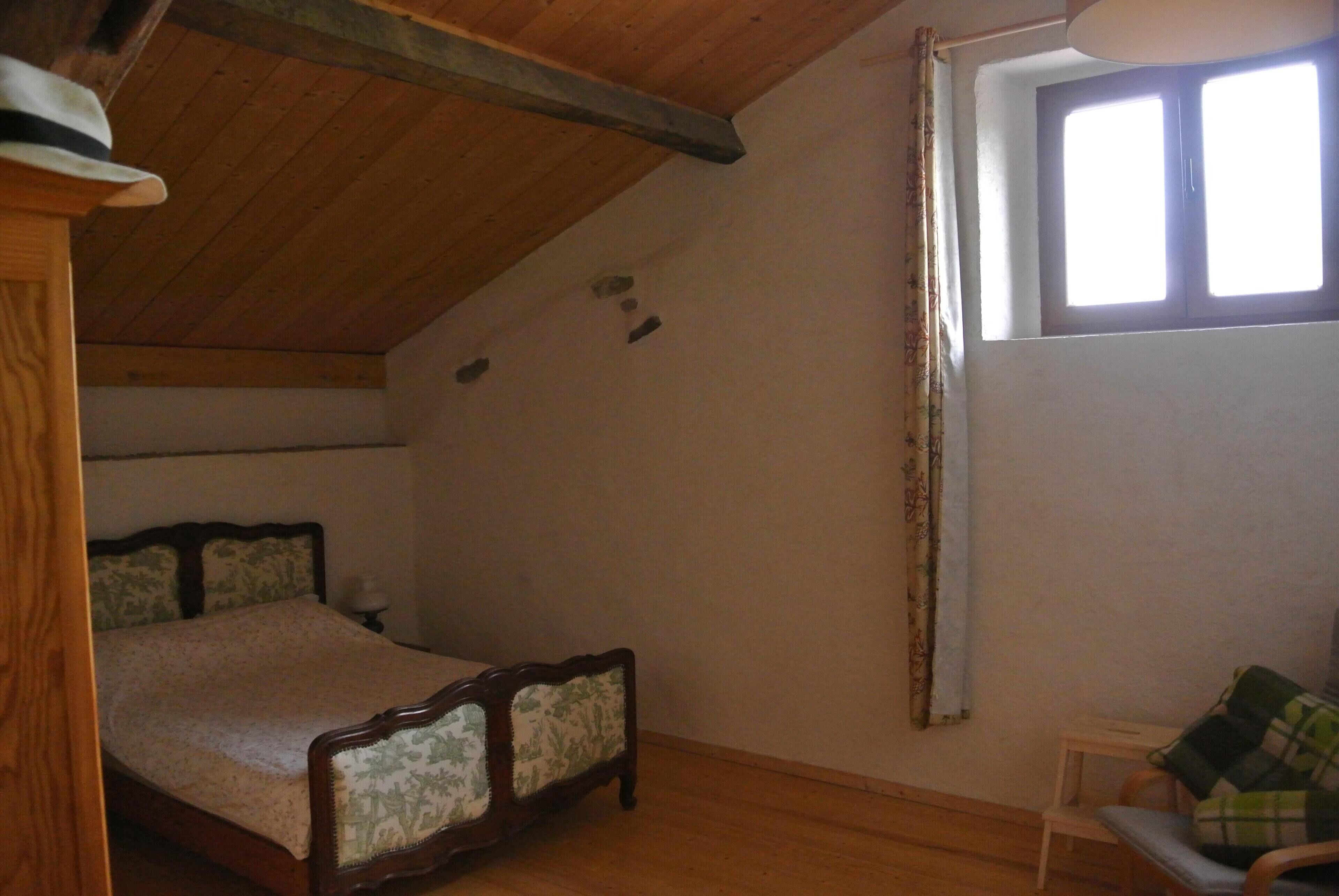 Ein Schlafzimmer mit Holzbalkendecke, einem Bett mit Blumenmuster am Kopfteil, einem kleinen Tisch und einem Fenster mit Vorhängen.