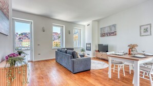A spacious living area with a grey sofa, wooden flooring, a dining table with chairs, and large windows offering a view of buildings outside. - The Perfect View Penthouse Design Apt. (Mali Losinj)