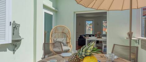 A dining area with a table set for two, featuring a plate with a pineapple, a melon, and a bowl of fruit. There are two wicker chairs, a white tablecloth, and a large umbrella providing shade.