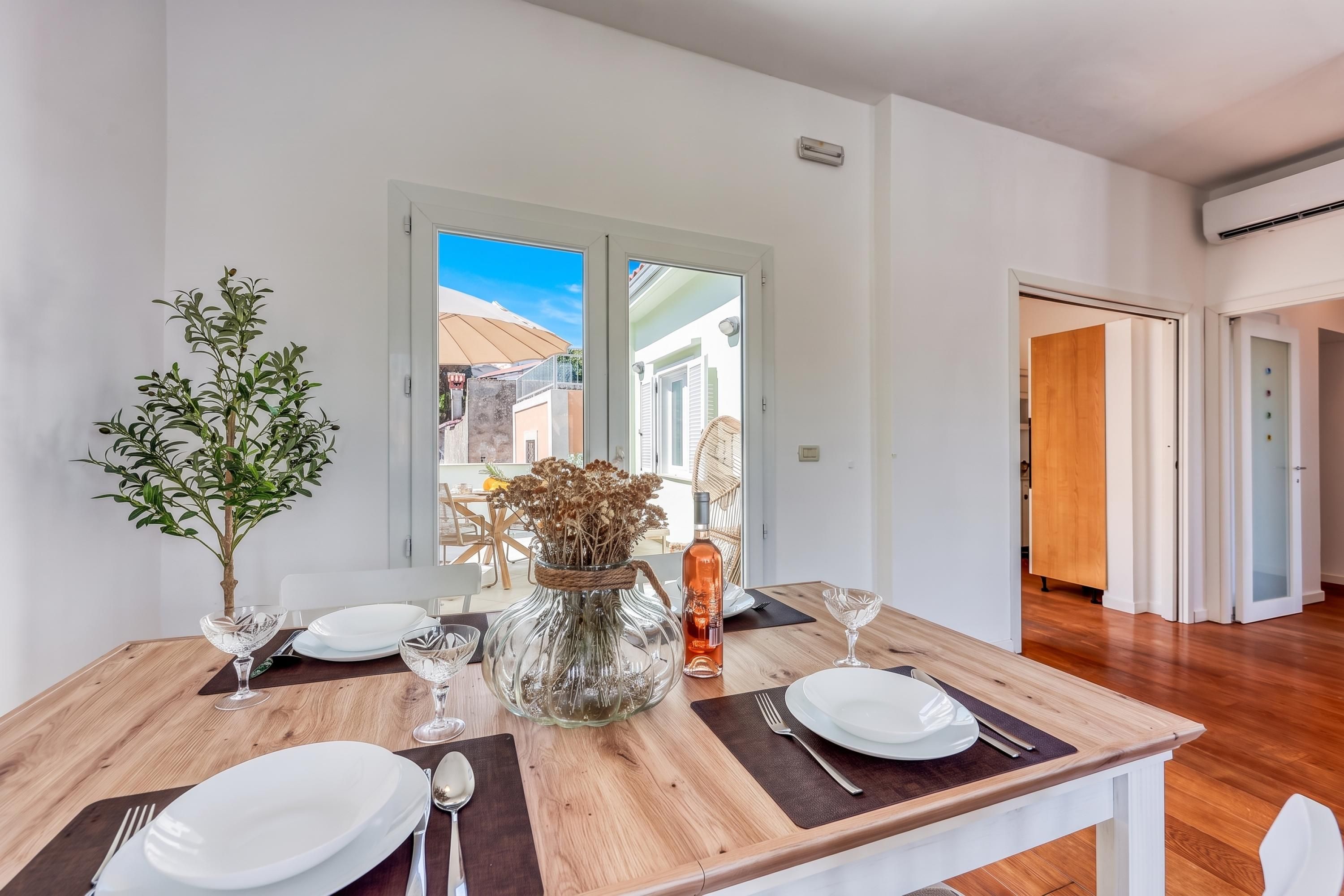 A dining area with a wooden table set for four, a potted plant, and a vase with dried flowers.