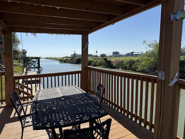 Une terrasse en bois avec une table et des chaises, d’où l’on a vue sur une étendue d’eau et sur des maisons au loin.