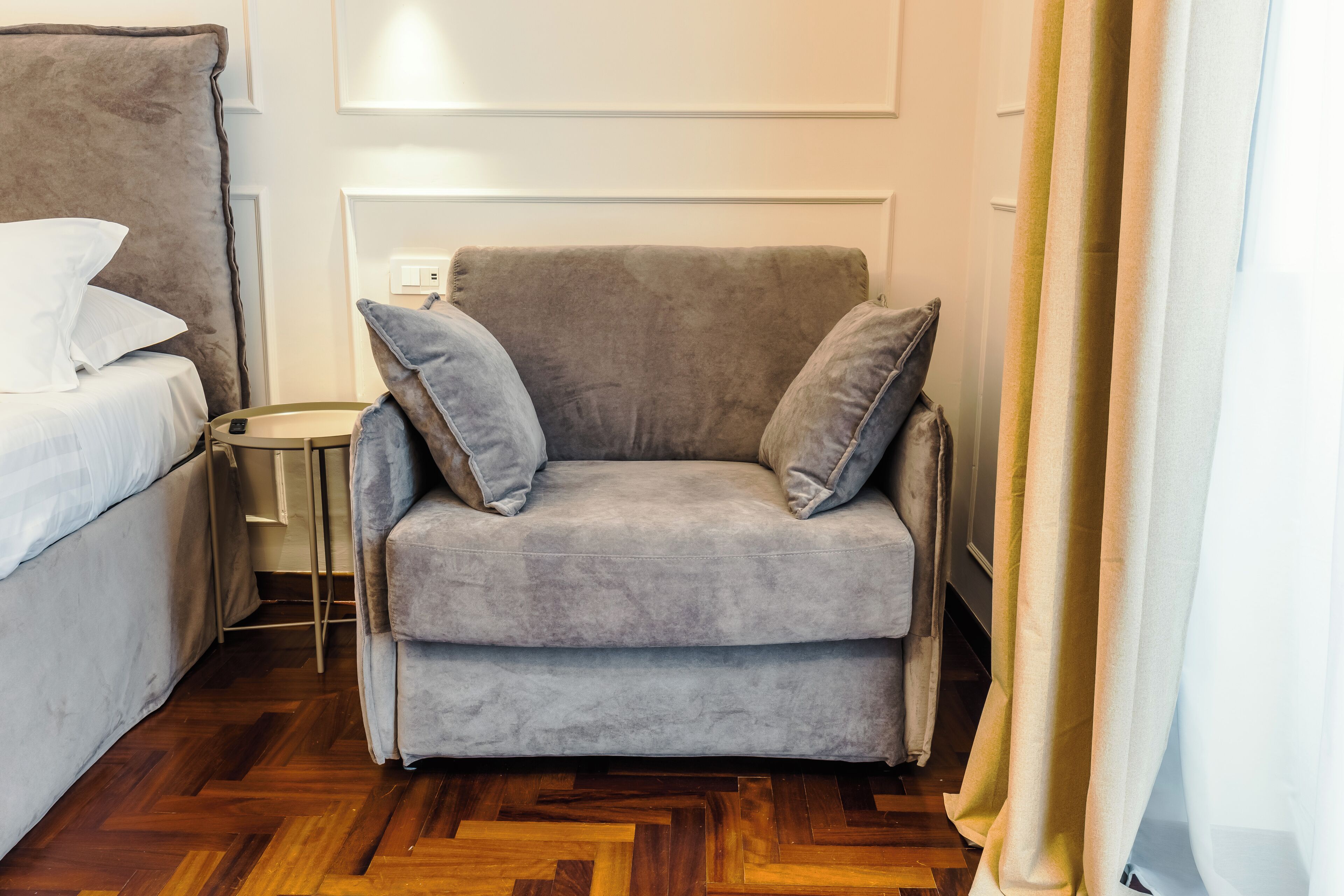 Een grijze bank met twee kussens, een klein rond tafeltje en een bed met wit beddengoed in een kamer met houten vloeren.