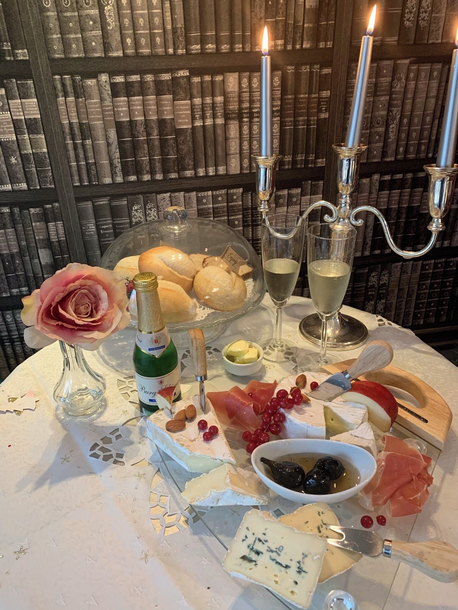 A table set with a variety of cheeses, fruits, a bottle of wine, and a rose in a vase, with candles and books in the background.