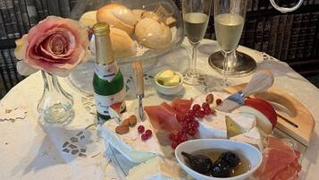 A table set with a variety of cheeses, fruits, a bottle of wine, and a rose in a vase, with candles and books in the background.