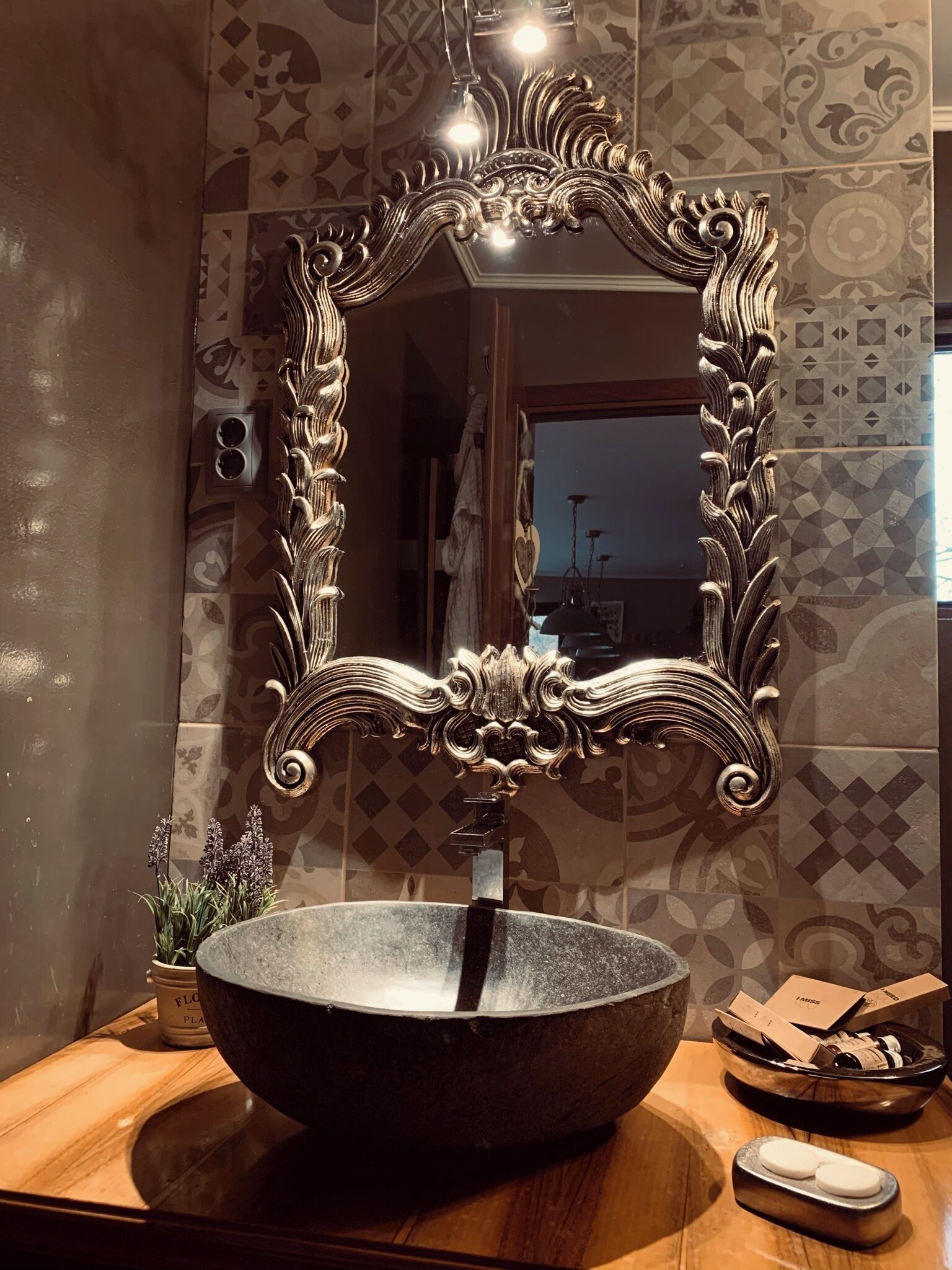 Un baño con un espejo grande y ornamentado, un lavabo de piedra y un mueble de tocador de madera.